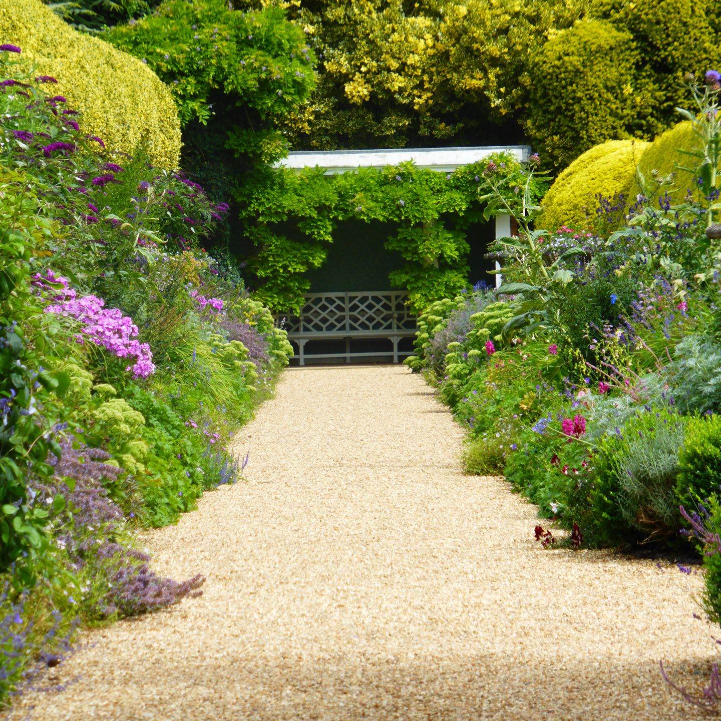Pathway through a colorful garden with a bench under a leafy pergola