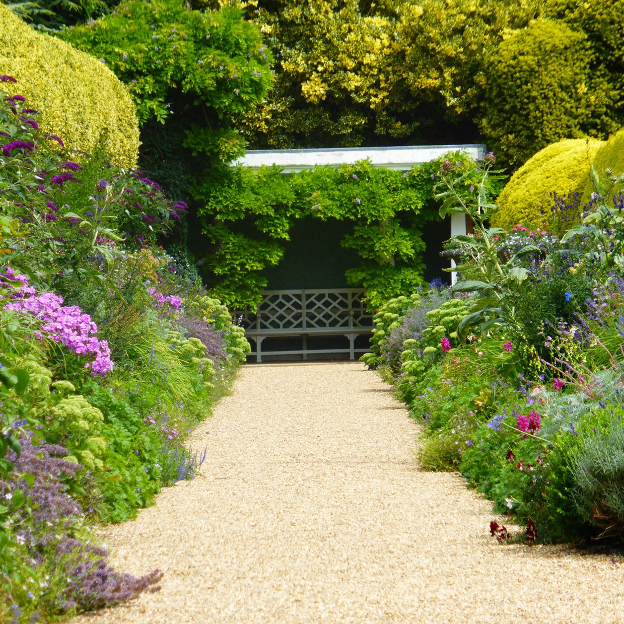 Pathway through a colorful garden with a bench under a leafy pergola