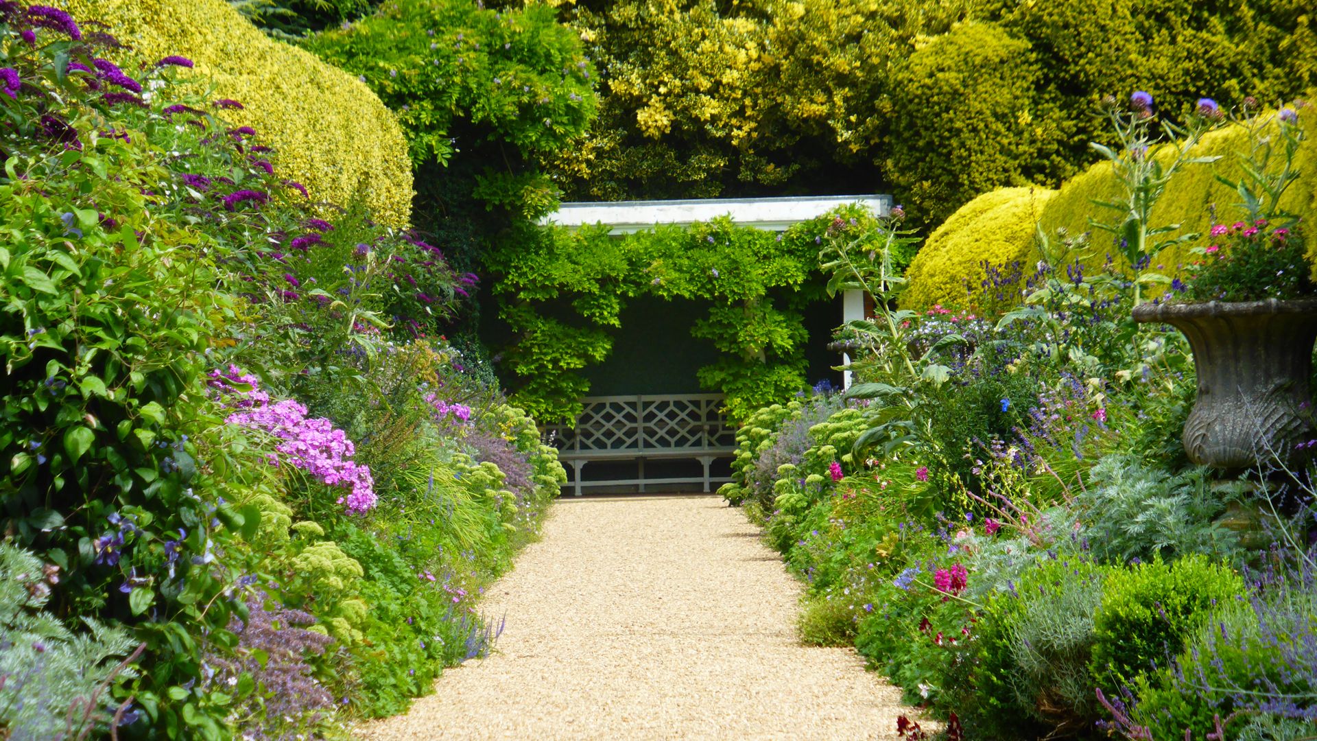 Pathway through a colorful garden with a bench under a leafy pergola