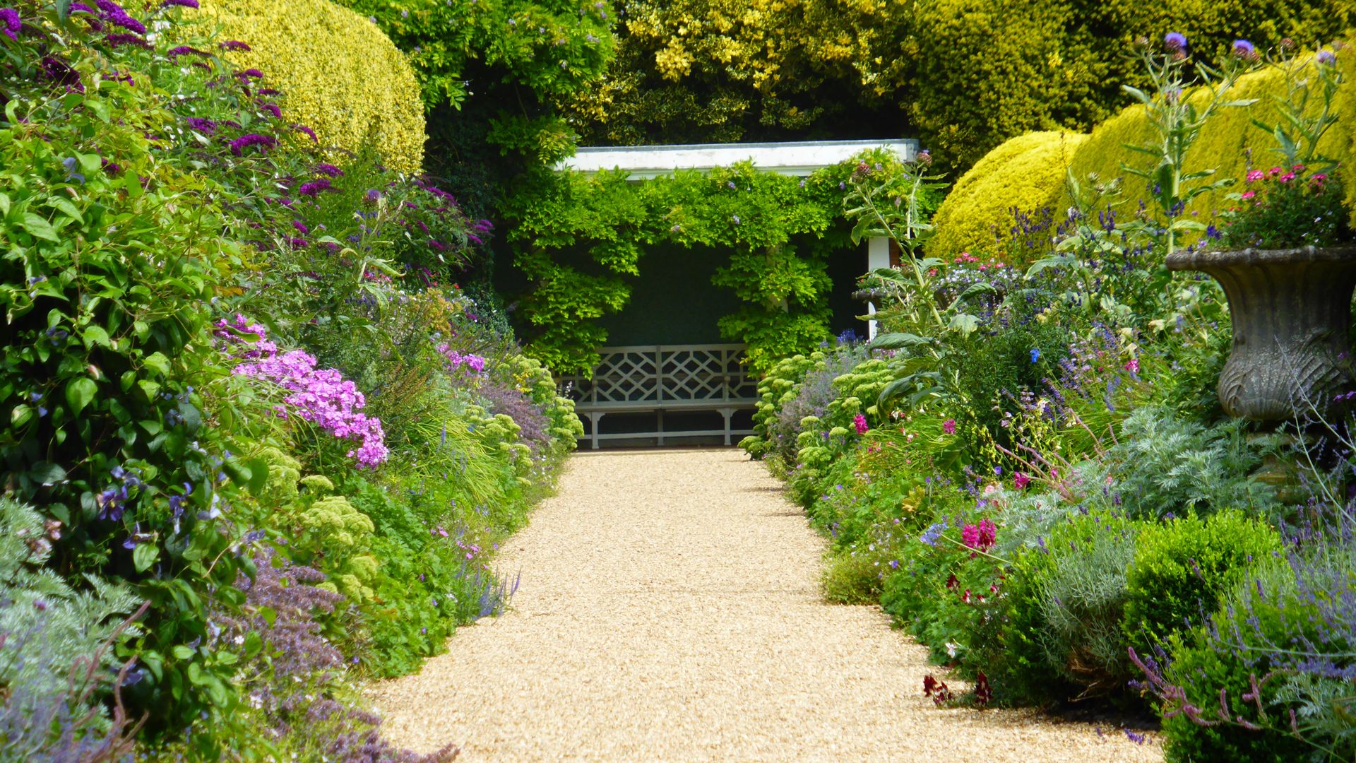 Pathway through a colorful garden with a bench under a leafy pergola