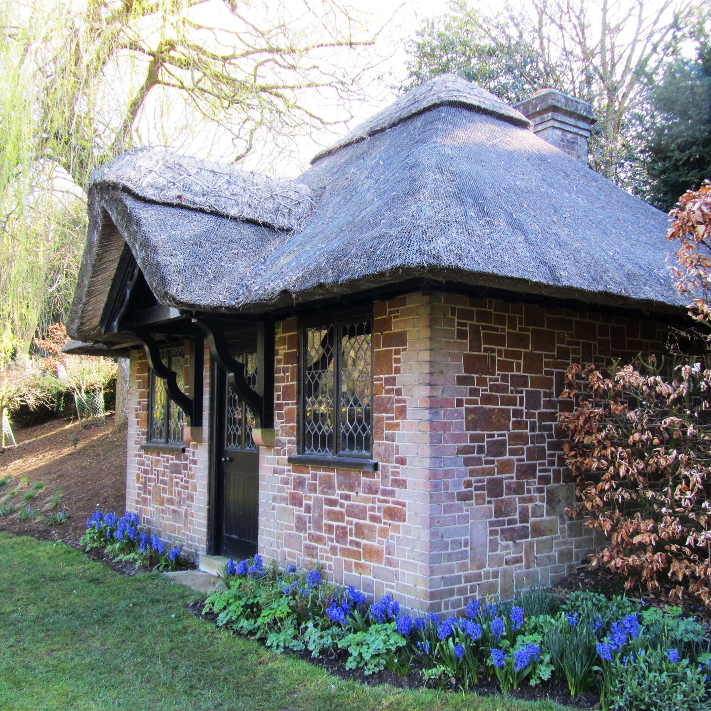 A quaint brick cottage with a thatched roof surrounded by garden flowers and trees.