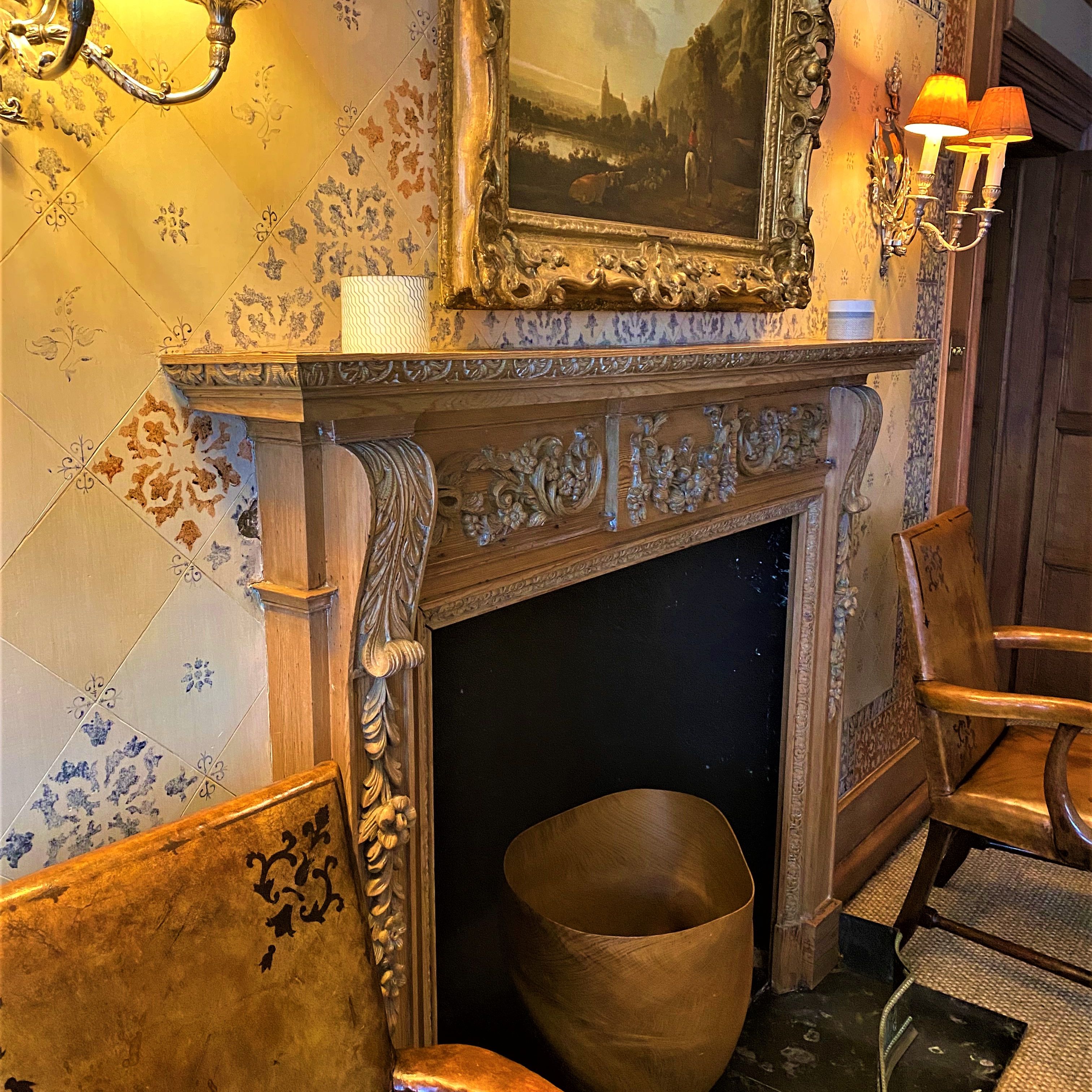Ornate fireplace with carved mantel, painting, wall lamps, and wooden chairs