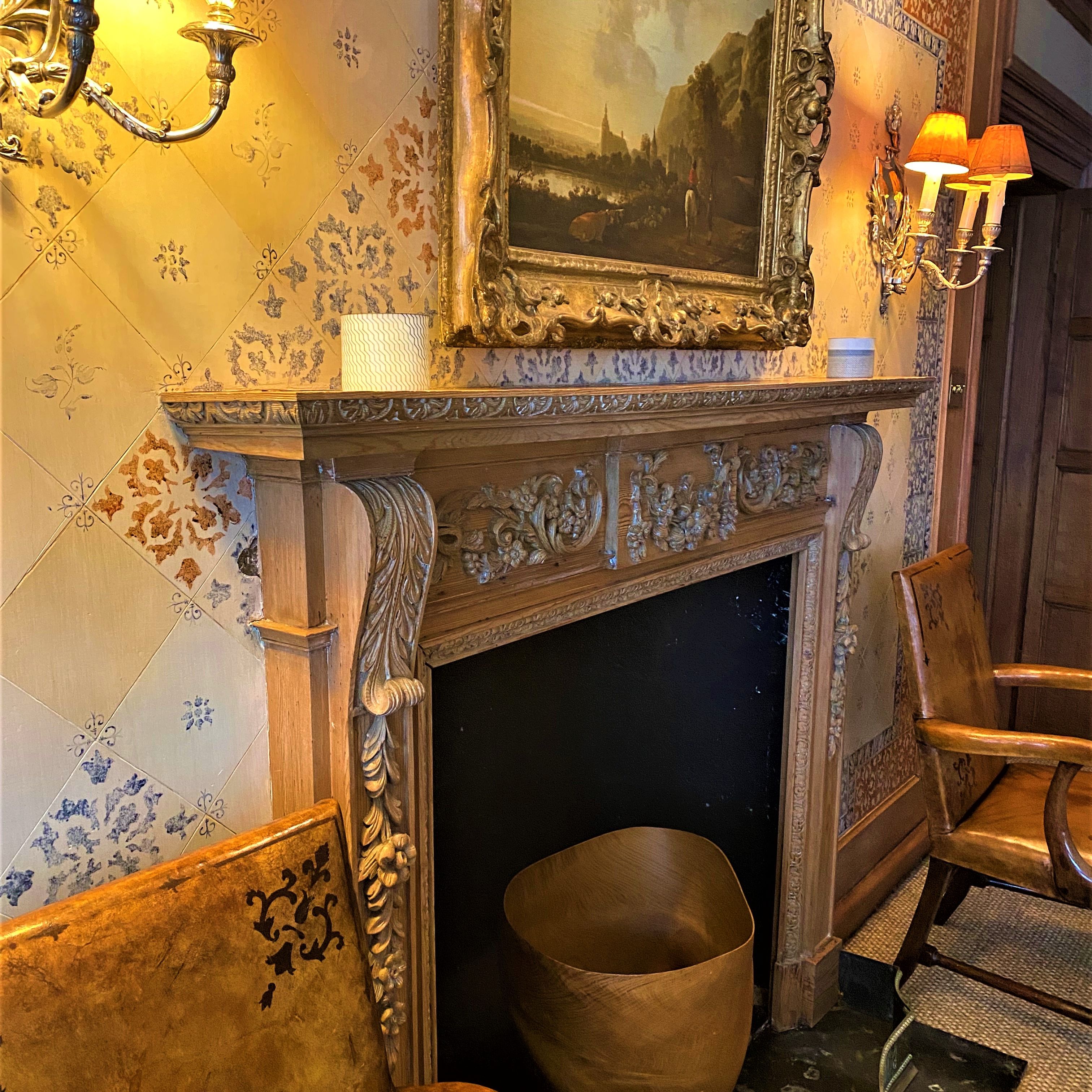 Ornate fireplace with carved mantel, painting, wall lamps, and wooden chairs