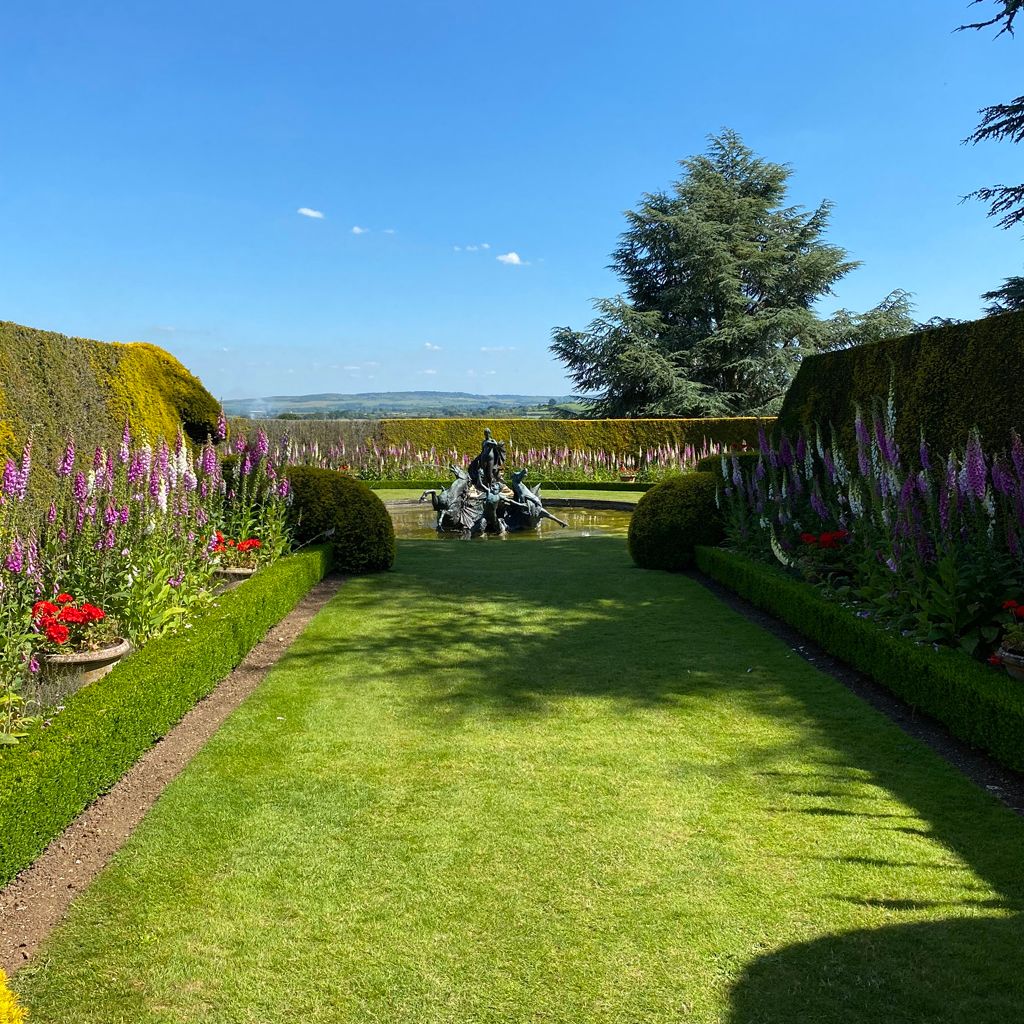Formal garden with manicured hedges, flowerbeds, and a statue fountain in the distance under a clear blue sky