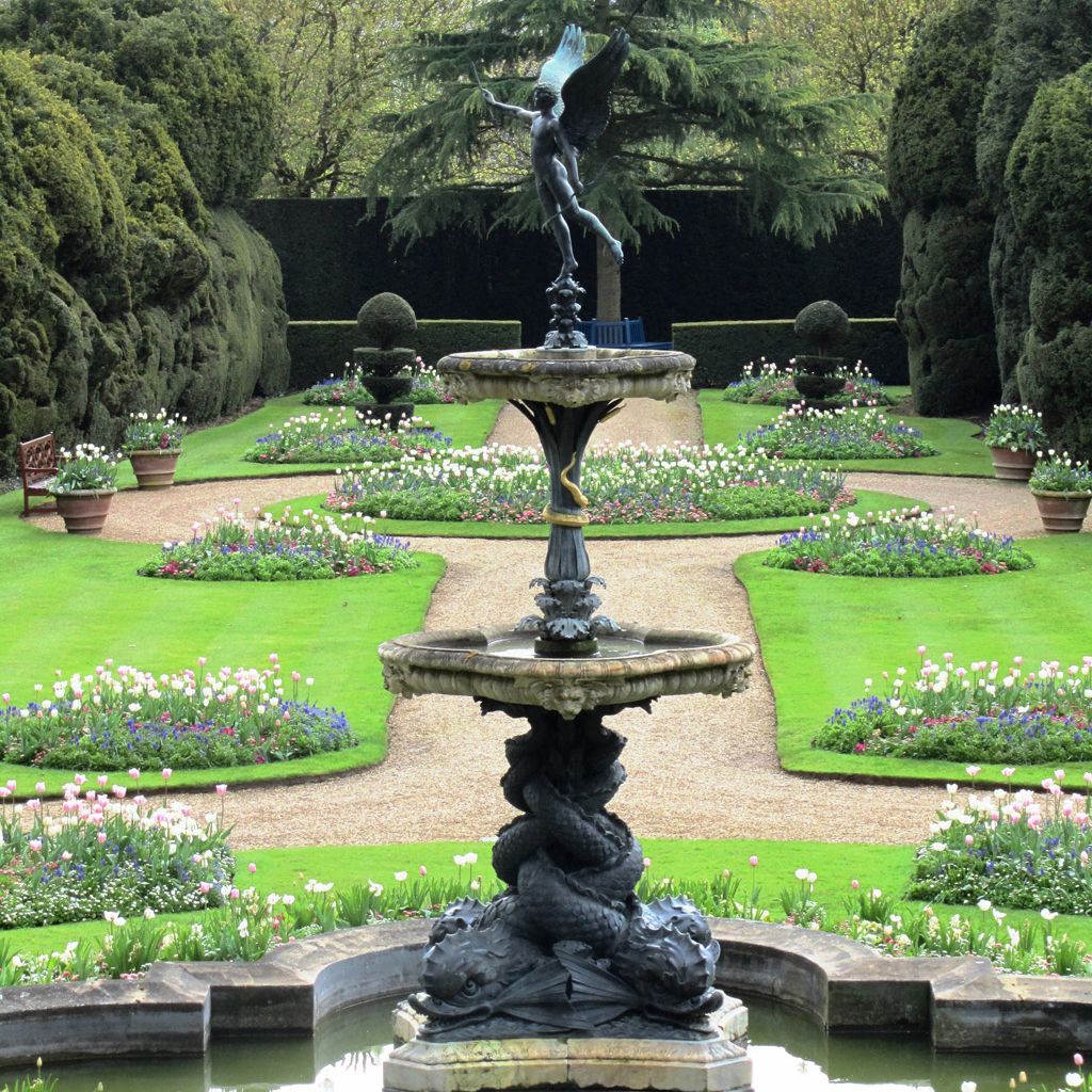 Elegant garden with a decorative fountain and a winged statue in the center, surrounded by manicured lawns and flower beds.