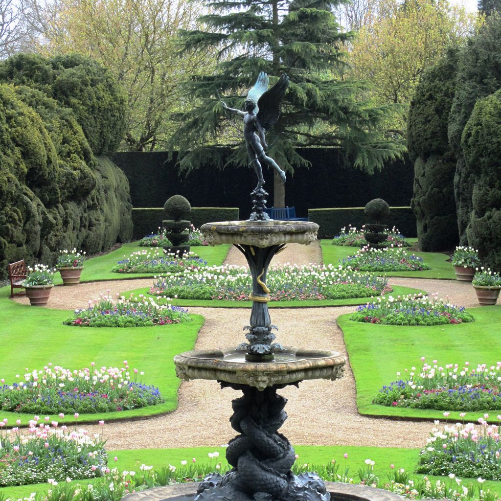 Elegant garden with a decorative fountain and a winged statue in the center, surrounded by manicured lawns and flower beds.