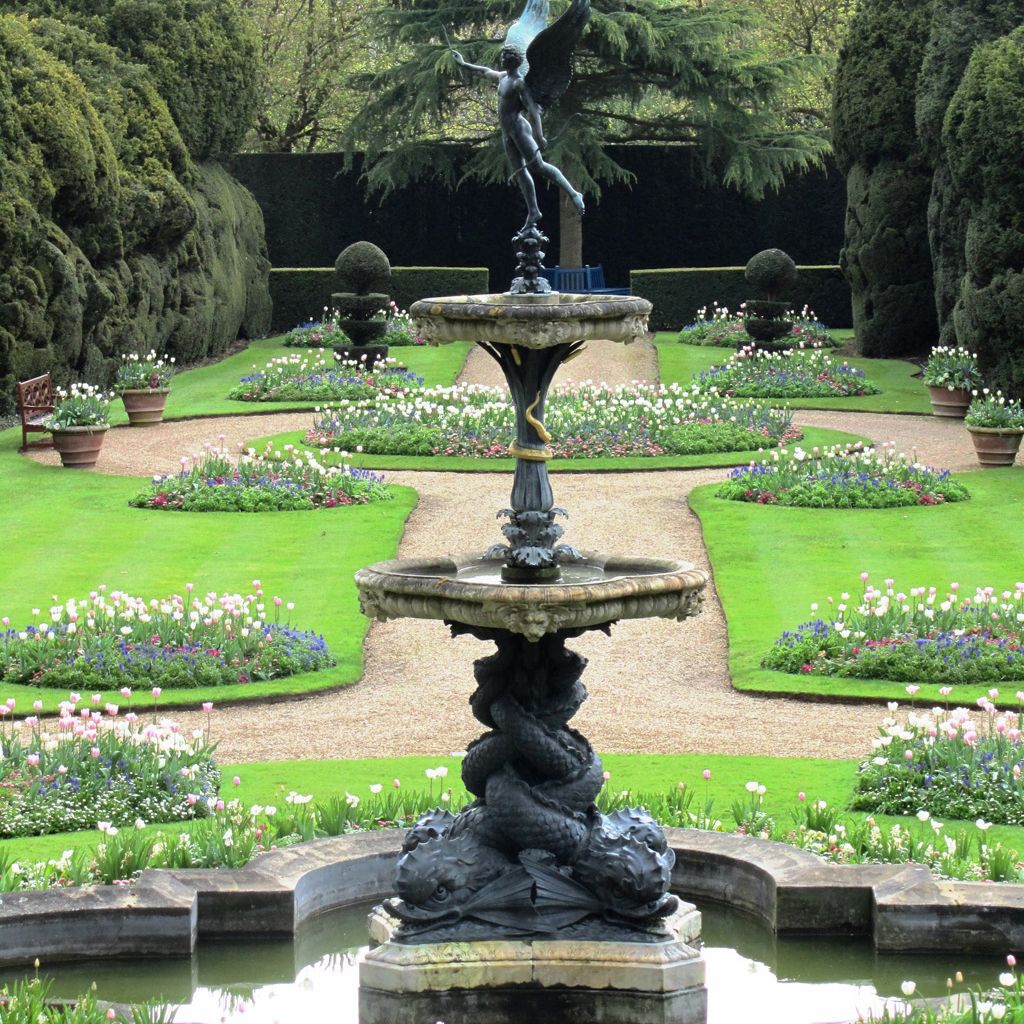 Elegant garden with a decorative fountain and a winged statue in the center, surrounded by manicured lawns and flower beds.