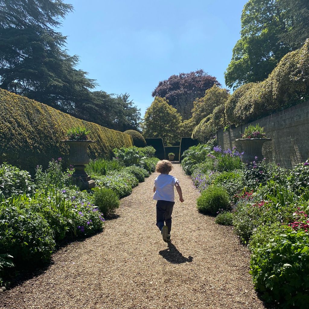 Child running down a gravel path through a lush, flower-filled garden on a sunny day