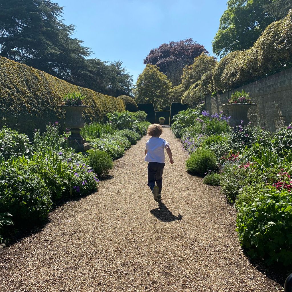 Child running down a gravel path through a lush, flower-filled garden on a sunny day