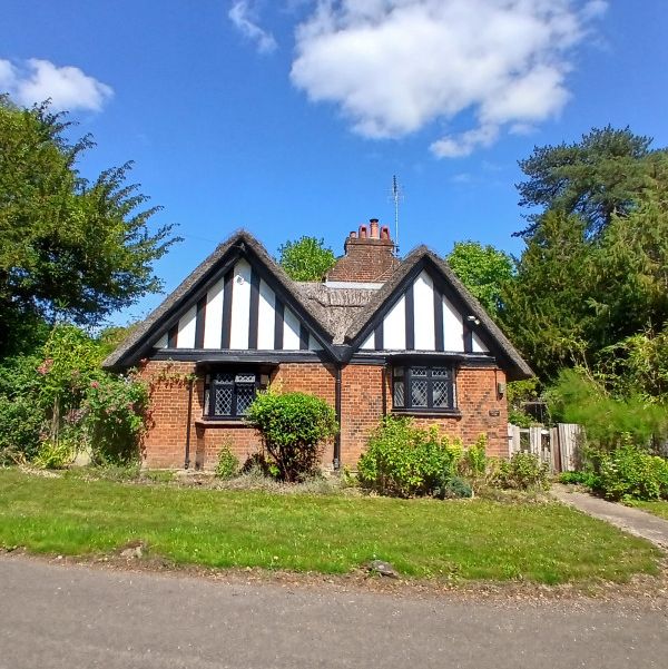A small, charming brick cottage with Tudor-style timber framing, surrounded by greenery and shrubs, under a blue sky with some clouds.