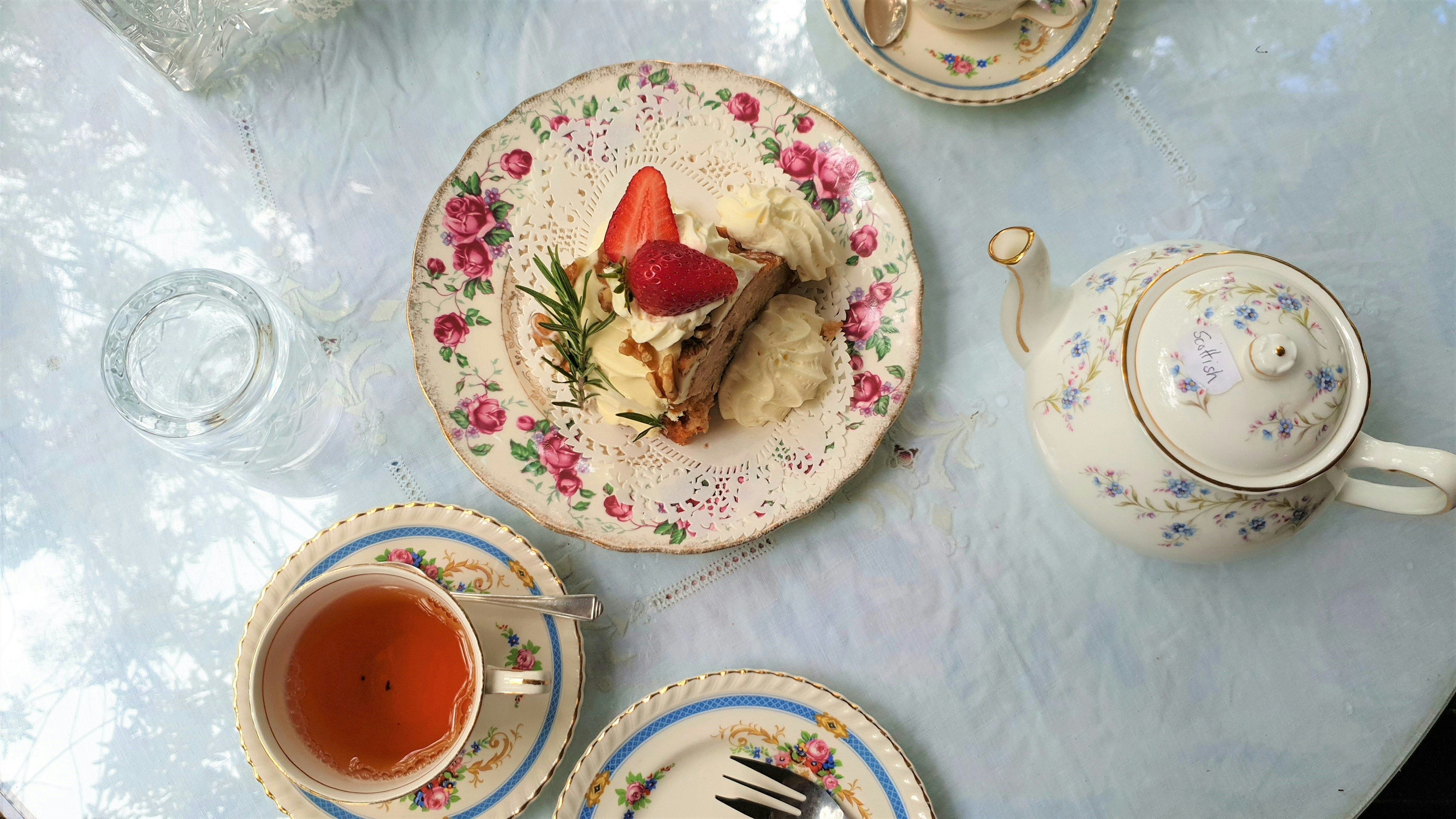 A floral-decorated tea set with a teapot, cups of tea, and a dessert with strawberries on a lace doily.