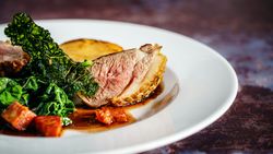 Close-up of a gourmet plated dish with roasted meat, sautéed greens, crispy kale, and sauce.