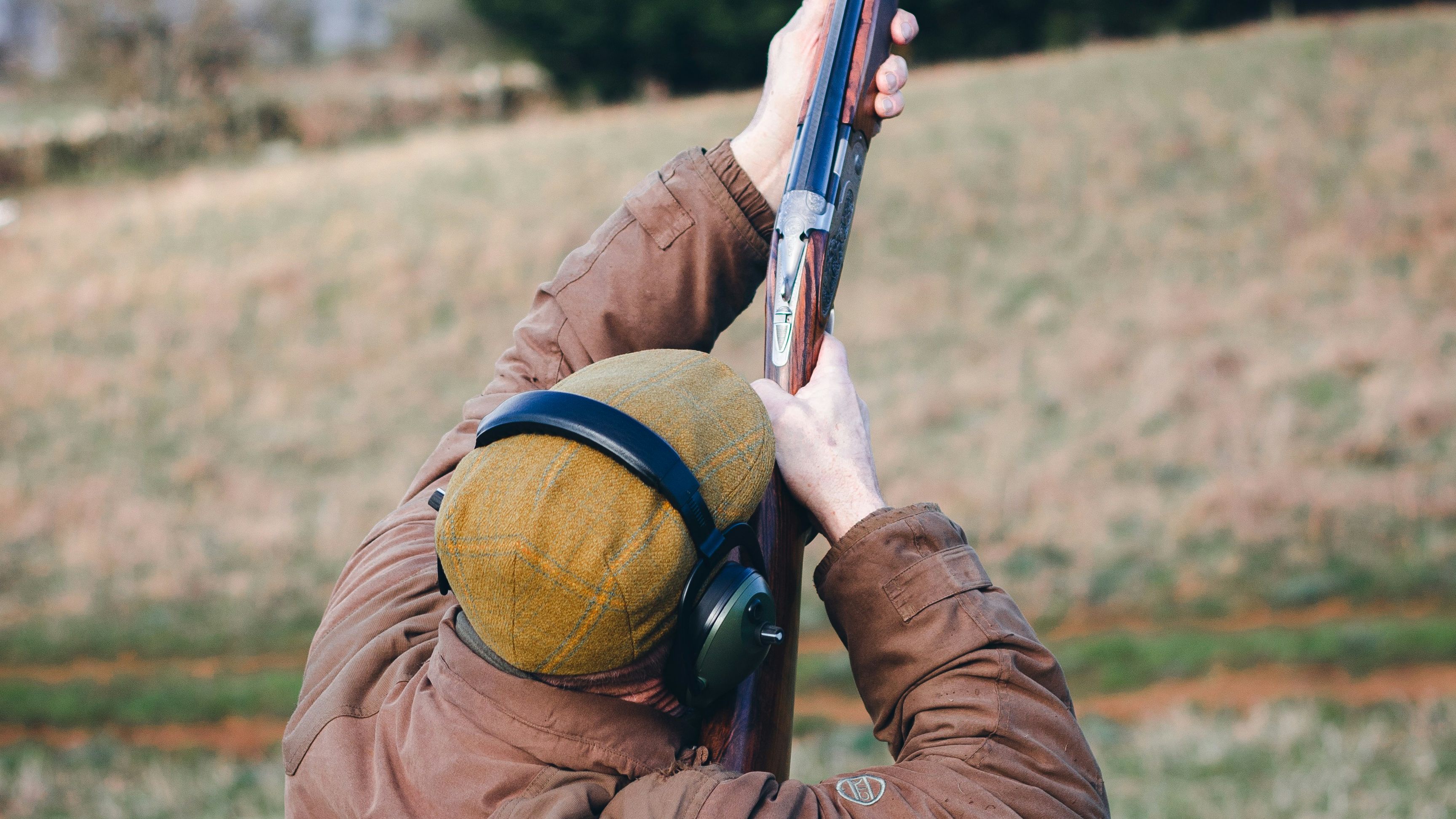 Person in brown jacket and earmuffs aiming a shotgun upwards outdoors