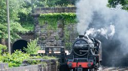 A black steam locomotive train with the number 5428 emerging from a stone tunnel surrounded by green foliage and smoke.