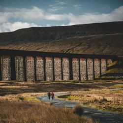 Two people walking on a path beside a large stone viaduct in a rural, mountainous landscape.