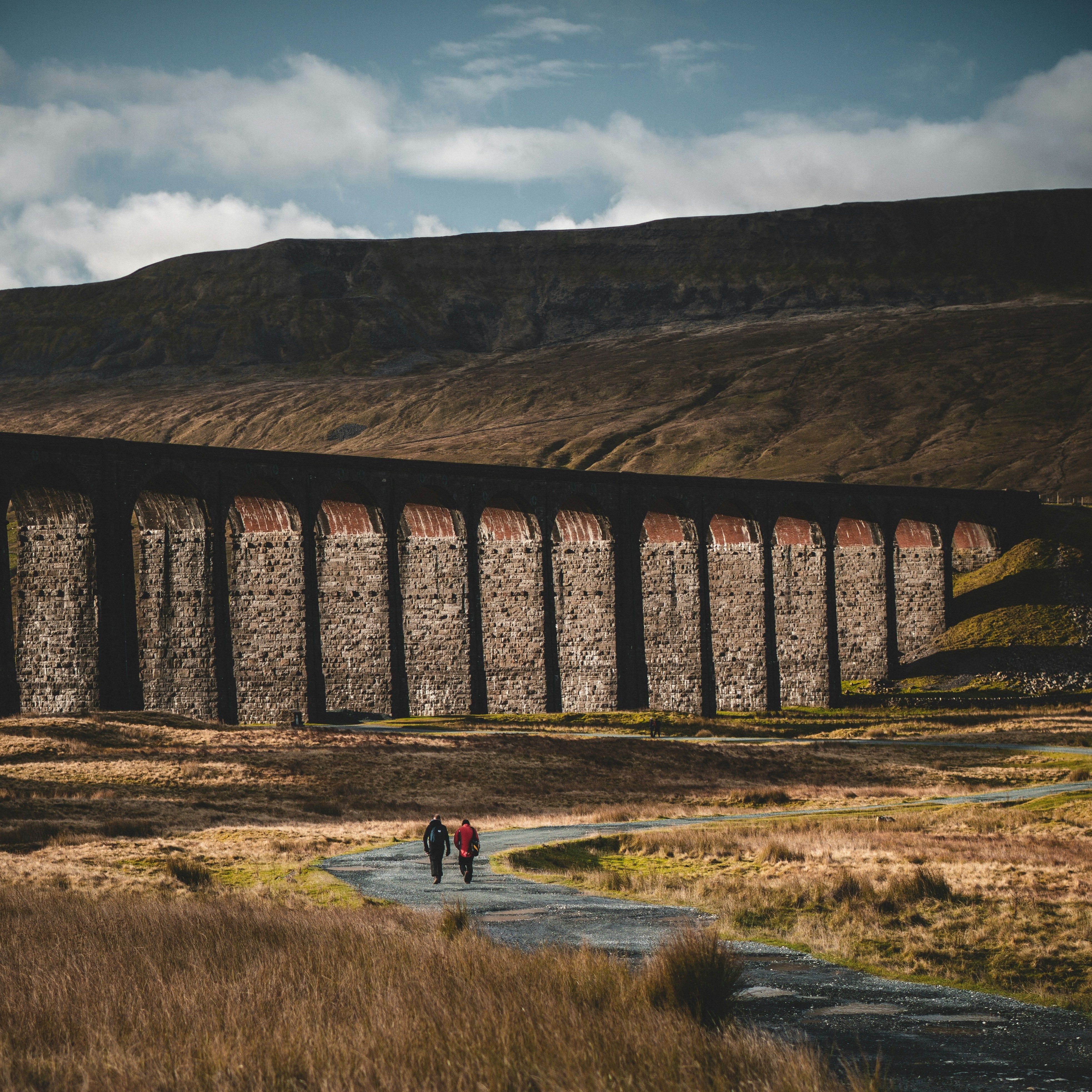 Two people walking on a path beside a large stone viaduct in a rural, mountainous landscape.