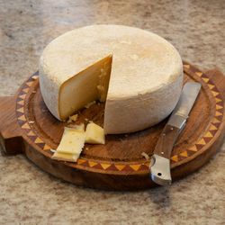 Whole wheel of cheese with a wedge cut out, slices of cheese, a knife, and a halved orange on a patterned wooden board.
