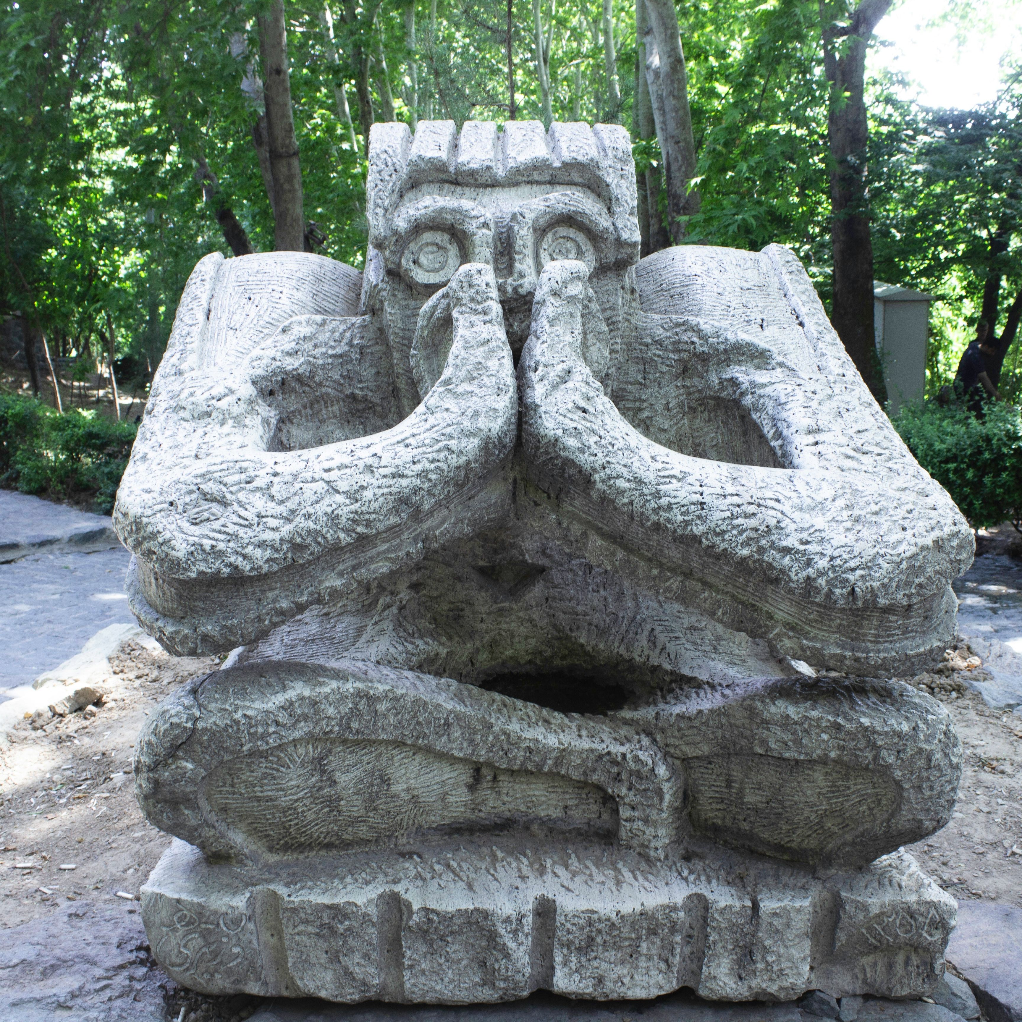 Large stone sculpture of a person sitting cross-legged with hands covering mouth and nose in an outdoor park setting.