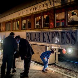People, including a child in pajamas, stand beside a vintage train marked 'Polar Express' and 'West Virginia' at night.