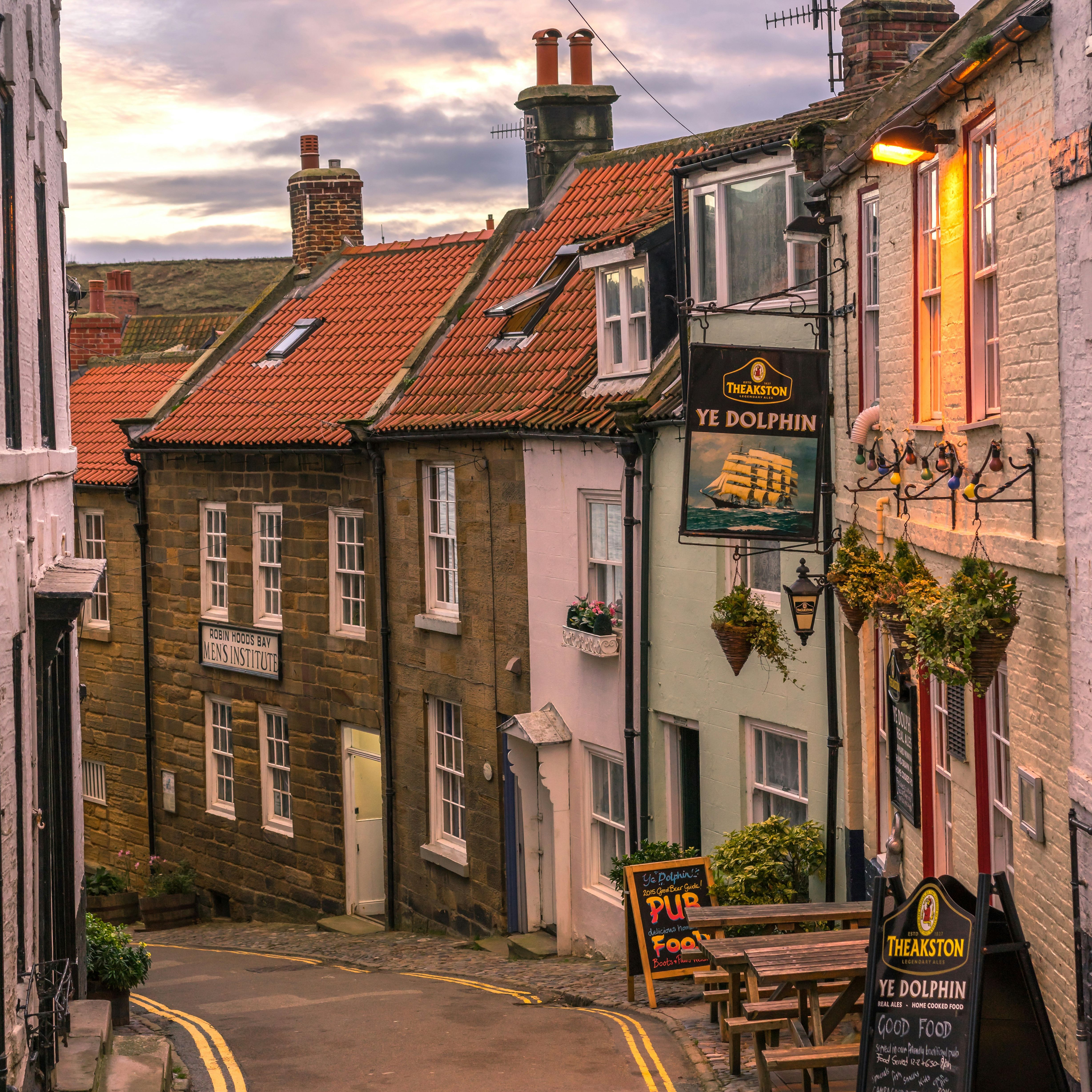 Quaint village street at sunset with cobblestone road, Ye Dolphin pub sign, and old stone houses with red tile roofs.