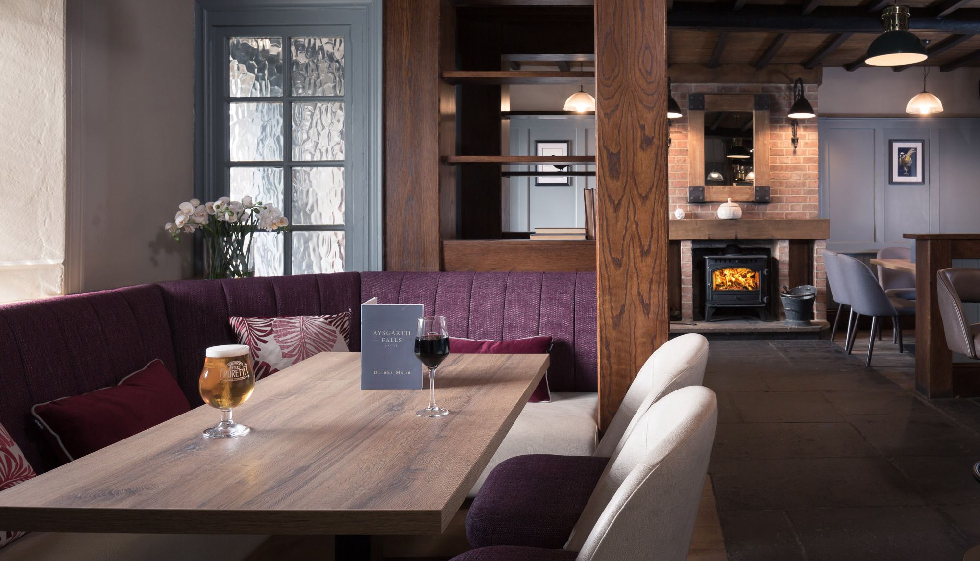 Cozy hotel lounge with wooden furnishings, a purple cushioned booth, drinks on the table, and a wood-burning fireplace.
