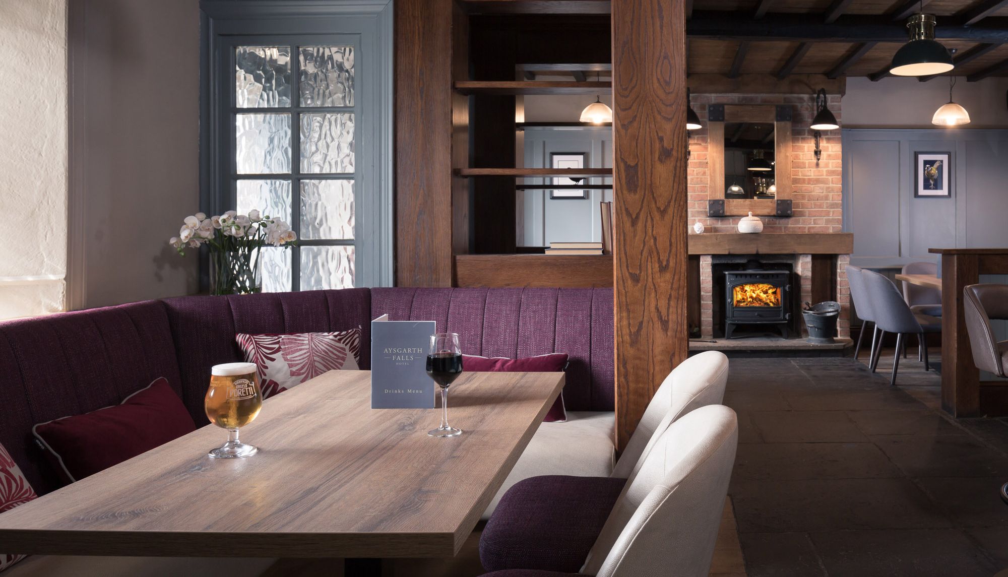 Cozy hotel lounge with wooden furnishings, a purple cushioned booth, drinks on the table, and a wood-burning fireplace.