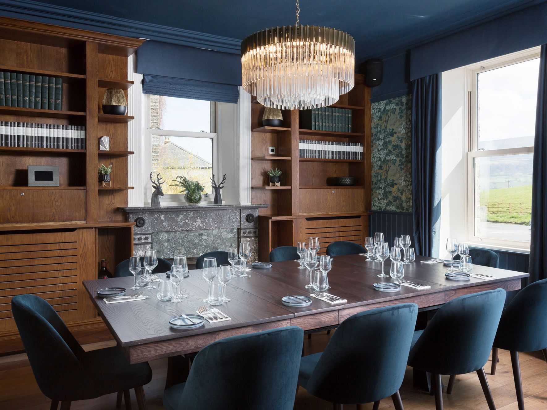 Elegant dining room with a large wooden table, blue velvet chairs, a fireplace, bookshelves, and a crystal chandelier