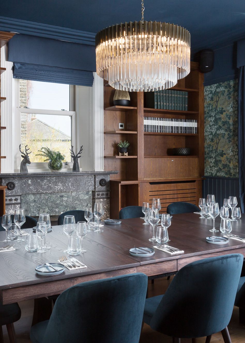 Elegant dining room with a large wooden table, blue velvet chairs, a fireplace, bookshelves, and a crystal chandelier