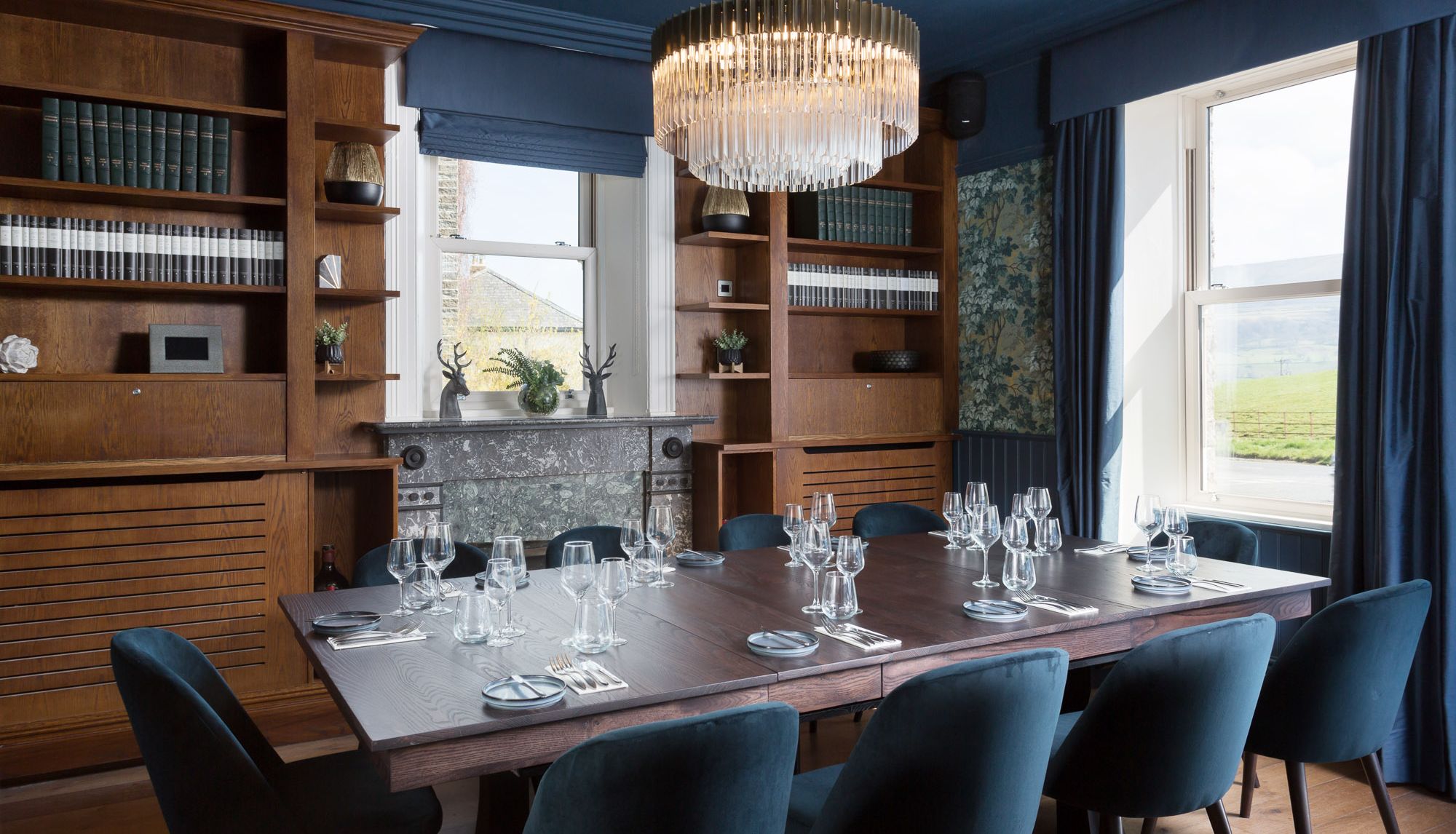 Elegant dining room with a large wooden table, blue velvet chairs, a fireplace, bookshelves, and a crystal chandelier