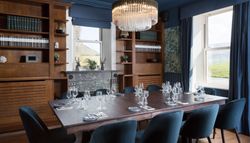 Elegant dining room with a large wooden table, blue velvet chairs, a fireplace, bookshelves, and a crystal chandelier