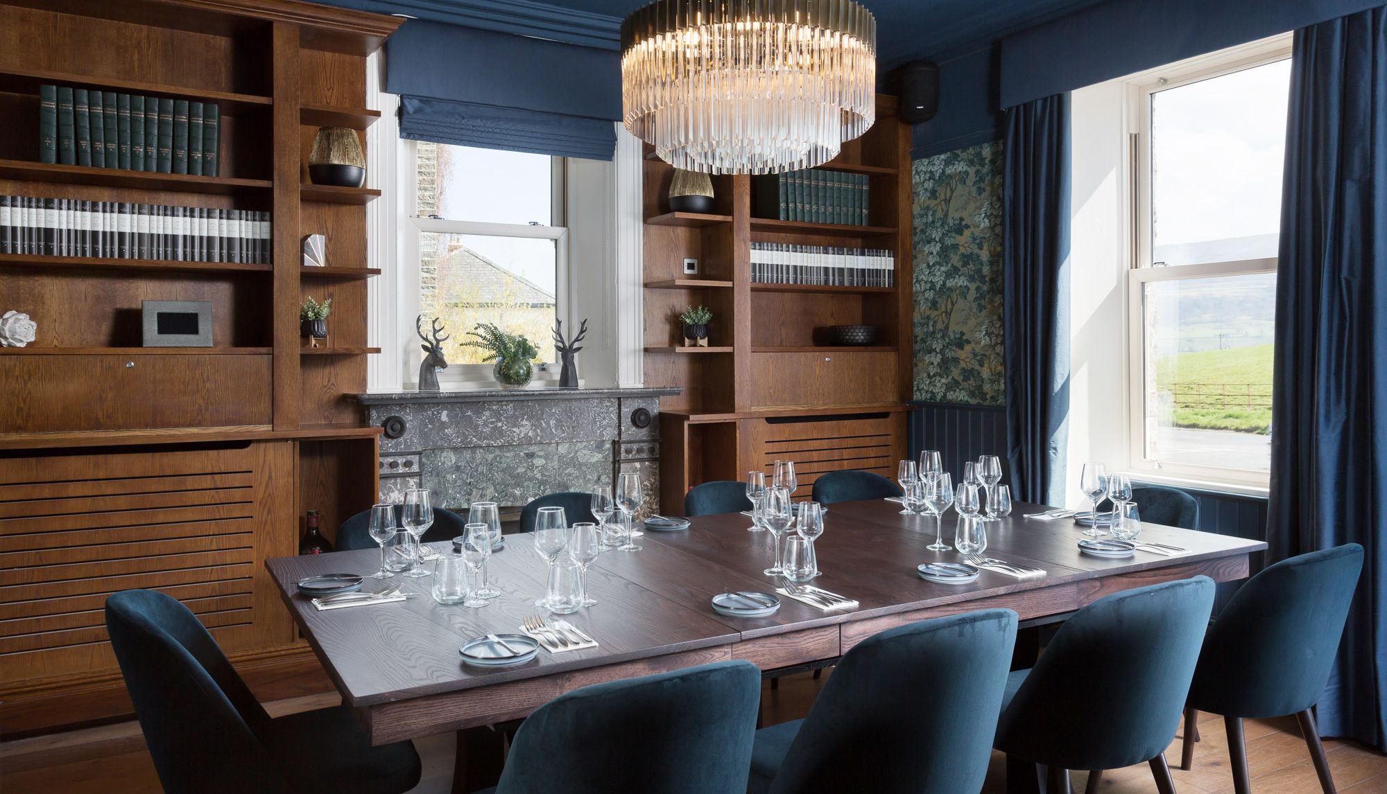 Elegant dining room with a large wooden table, blue velvet chairs, a fireplace, bookshelves, and a crystal chandelier