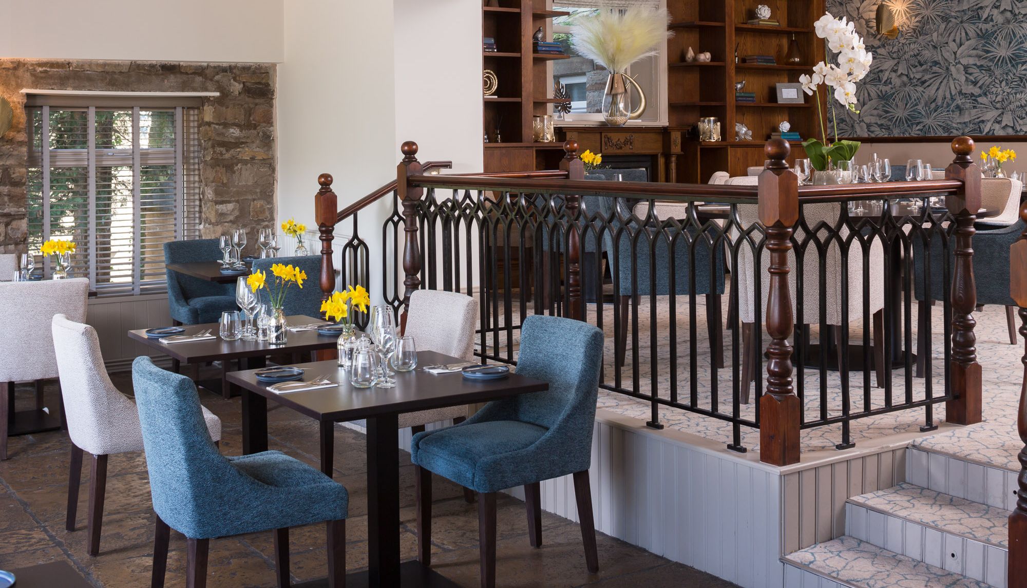 Modern restaurant dining area with blue and beige chairs, neatly set tables, yellow flowers, and a decorative railing separating two levels.