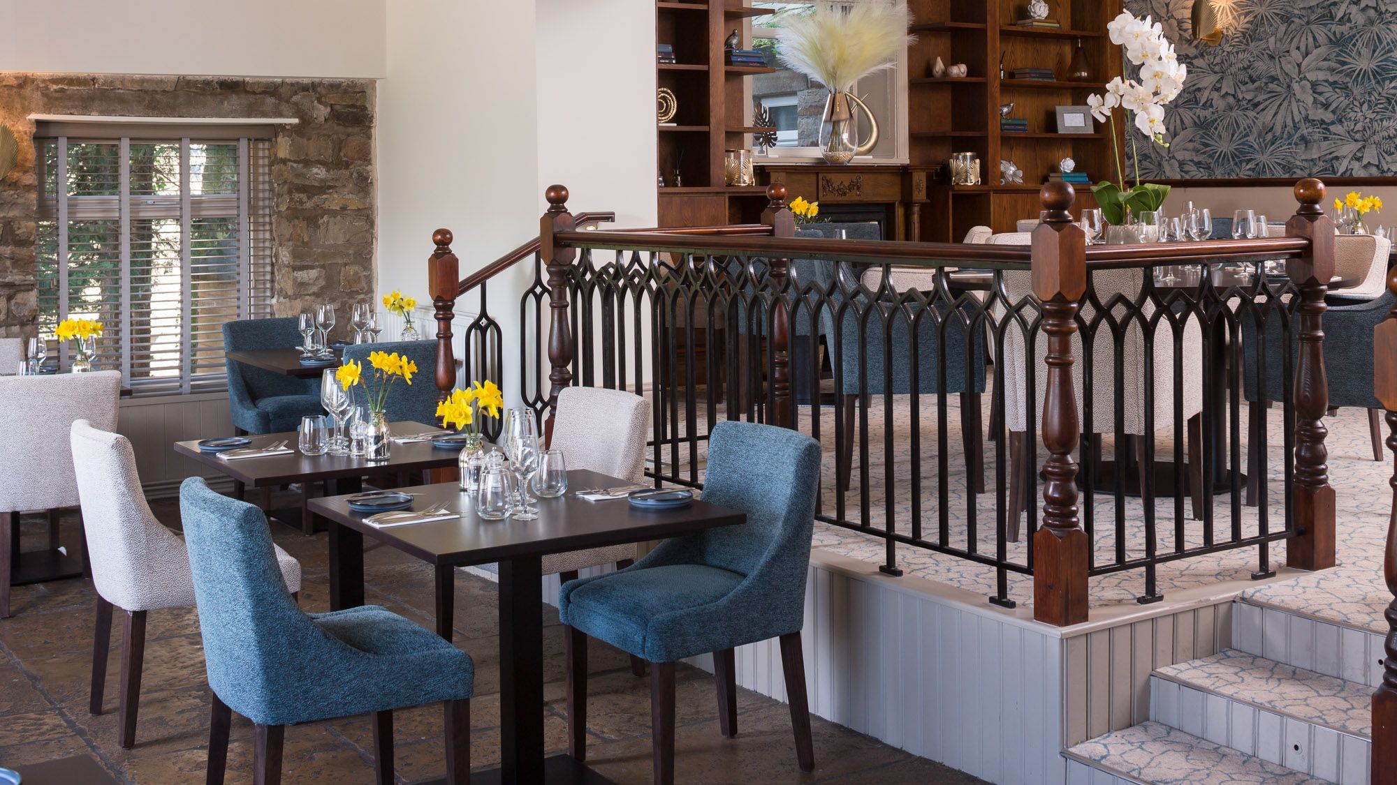 Modern restaurant dining area with blue and beige chairs, neatly set tables, yellow flowers, and a decorative railing separating two levels.