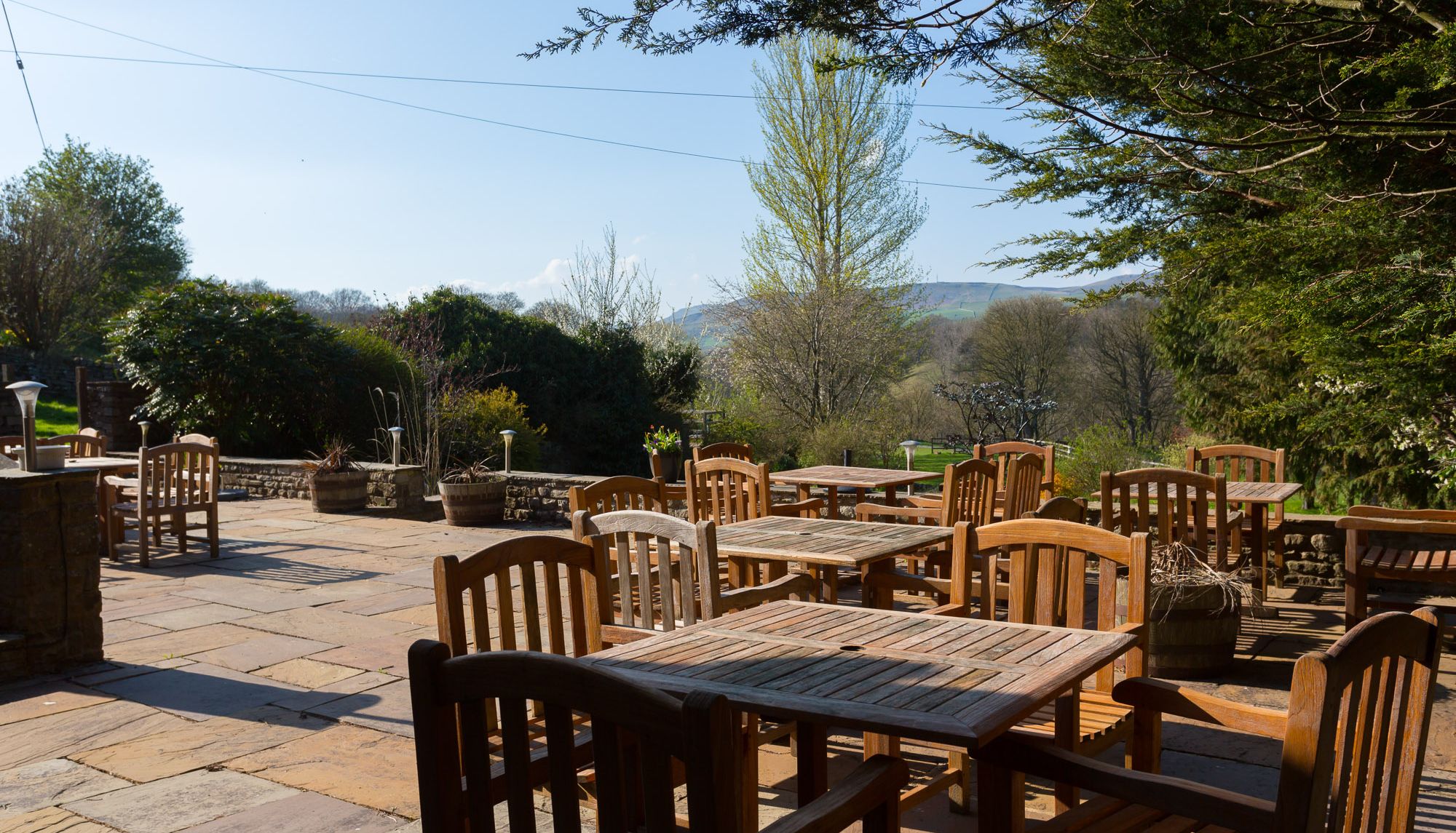 Outdoor seating area with wooden tables and chairs on a stone patio surrounded by greenery and trees.