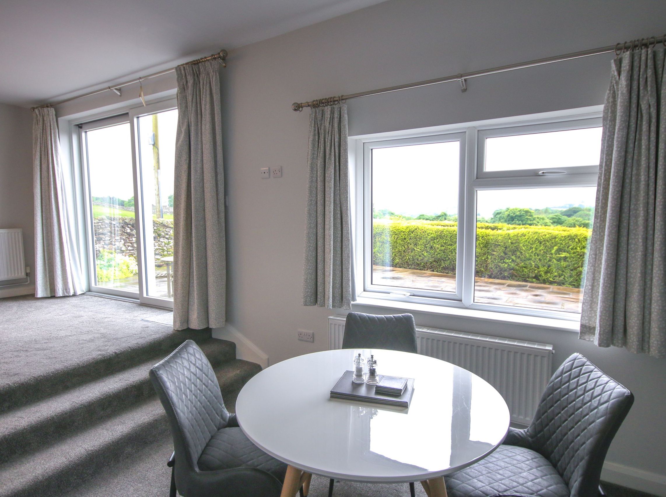 Modern cottage apartment interior with a round dining table, chairs, large window, and terrace view.