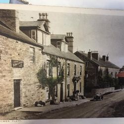 A vintage photograph of a small village street lined with old stone buildings and a car from the early 20th century.