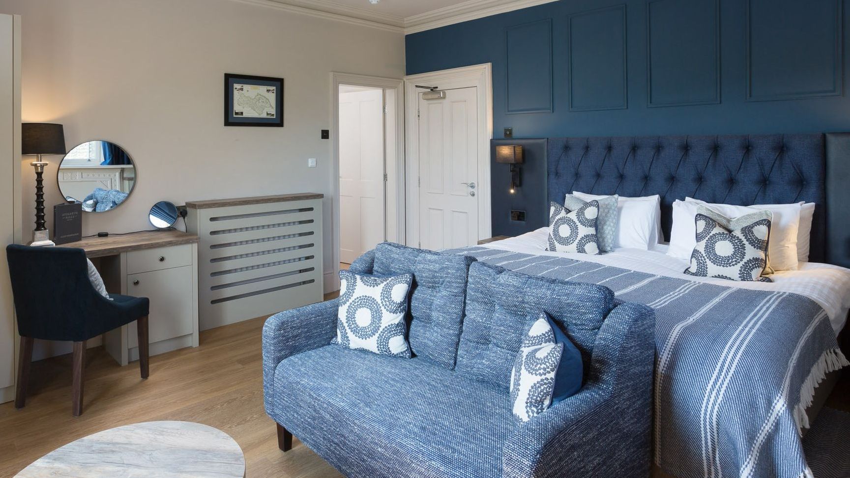 Modern hotel room with blue and white decor, a large bed, blue sofa, and work desk