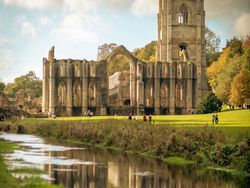 Ruins of a large stone abbey beside a river with reflection, surrounded by grass and trees under a partly cloudy sky