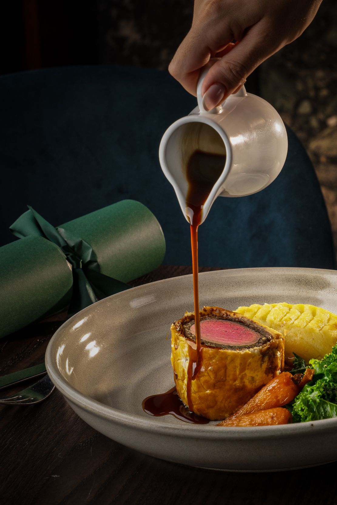A gourmet meal featuring beef Wellington with vegetables and mashed potatoes, with gravy being poured, alongside a glass of sparkling wine.
