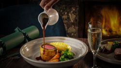 A gourmet meal featuring beef Wellington with vegetables and mashed potatoes, with gravy being poured, alongside a glass of sparkling wine.