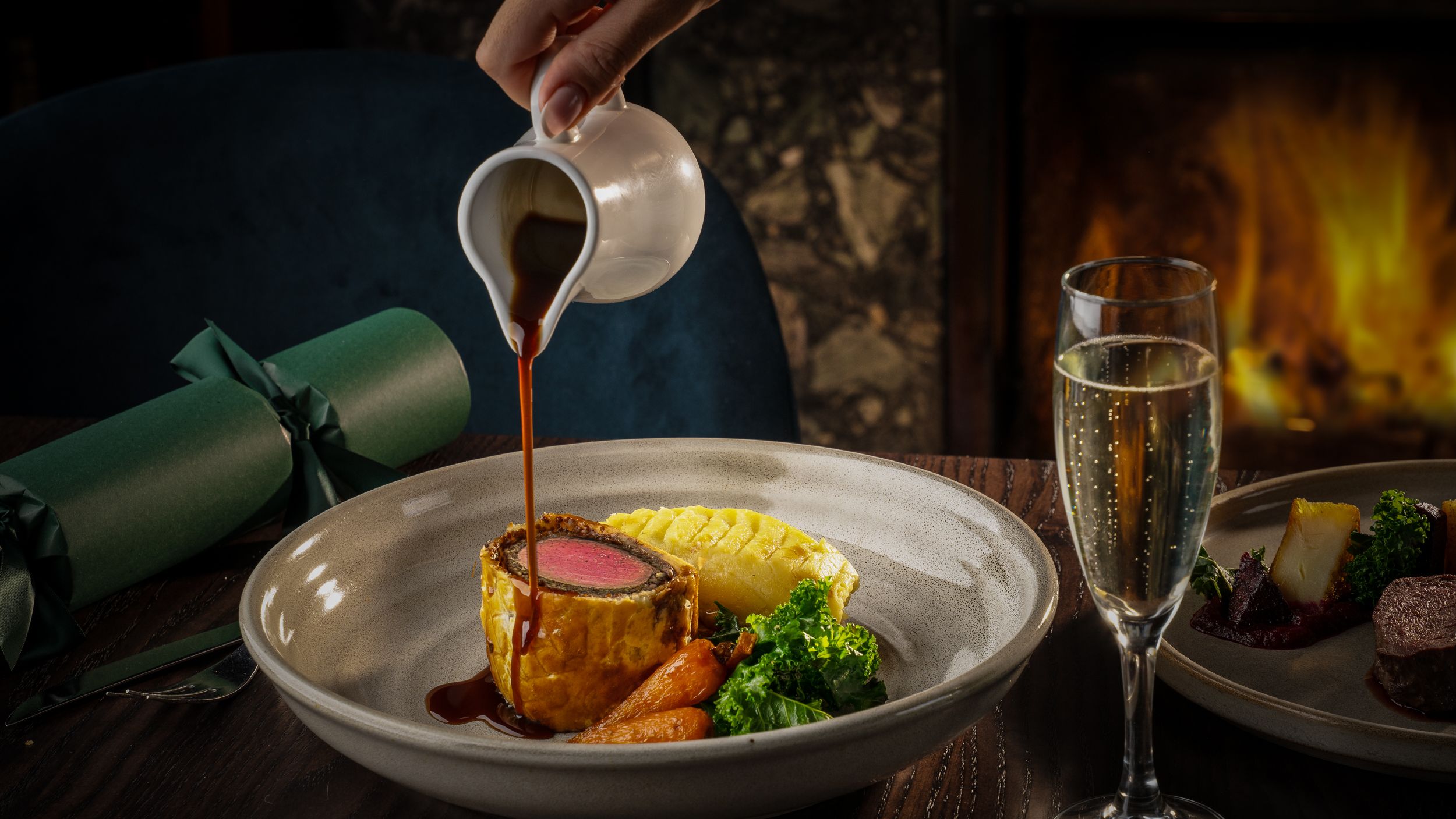 A gourmet meal featuring beef Wellington with vegetables and mashed potatoes, with gravy being poured, alongside a glass of sparkling wine.