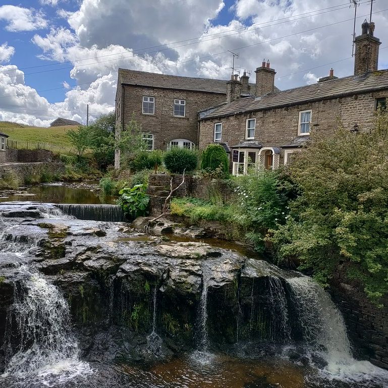 Stone houses beside a small waterfall and stream in a rural village setting under a partly cloudy sky.