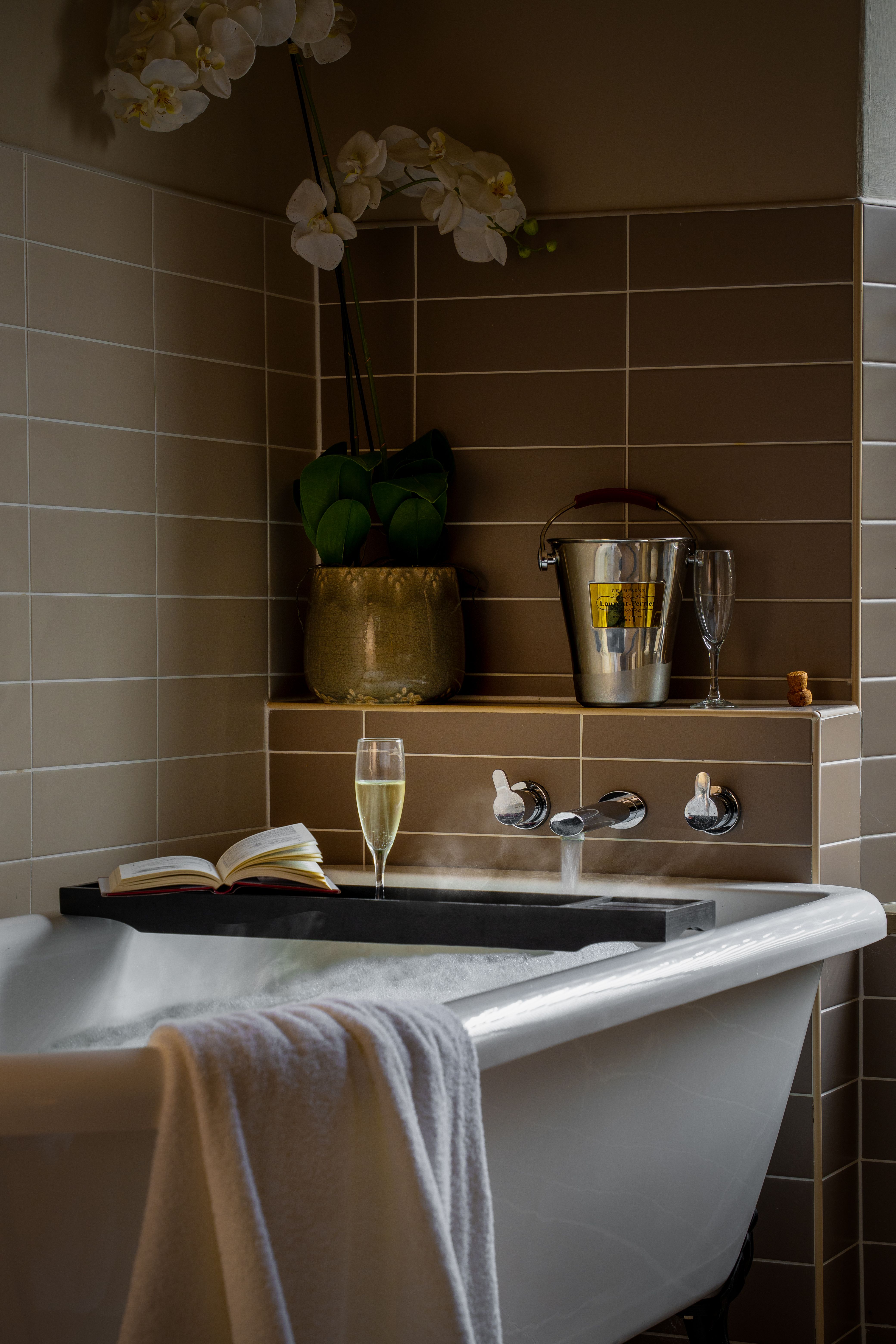 A luxurious bathtub with a towel draped over the edge, a book, a glass of champagne, and an ice bucket with a champagne bottle and glass on a tiled ledge.