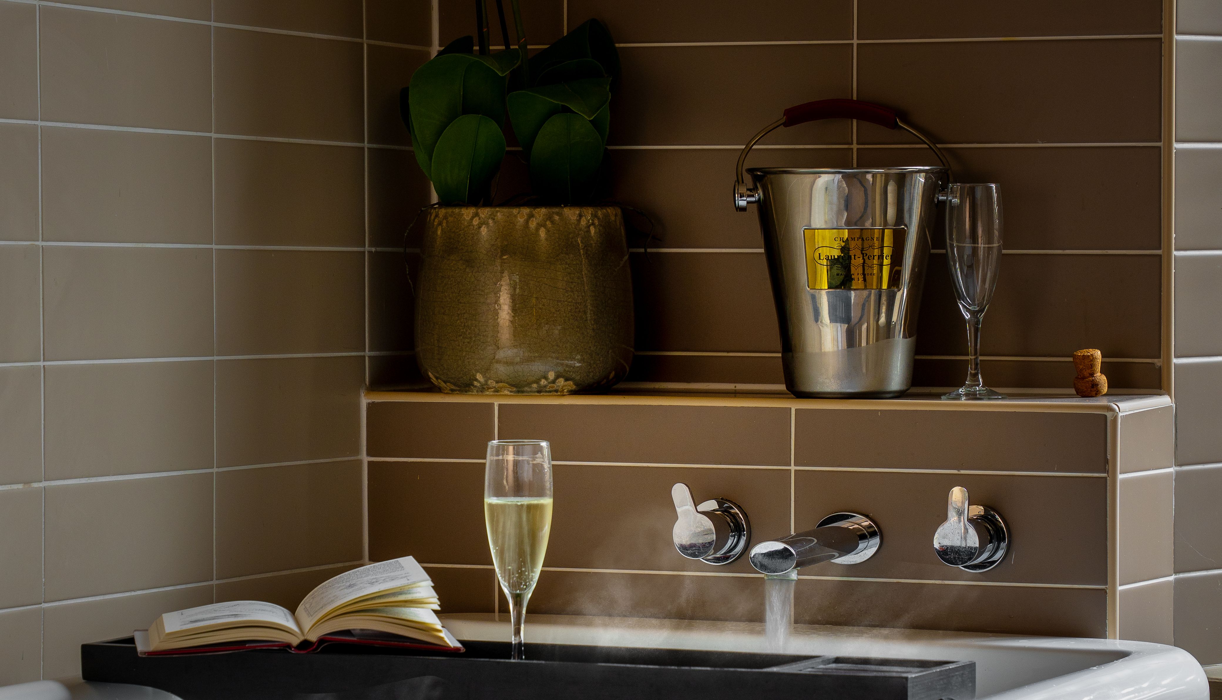 A luxurious bathtub with a towel draped over the edge, a book, a glass of champagne, and an ice bucket with a champagne bottle and glass on a tiled ledge.