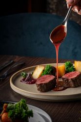 Hand pouring sauce over sliced beef tenderloin with vegetables and a glass of red wine on a dinner table.