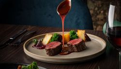 Hand pouring sauce over sliced beef tenderloin with vegetables and a glass of red wine on a dinner table.