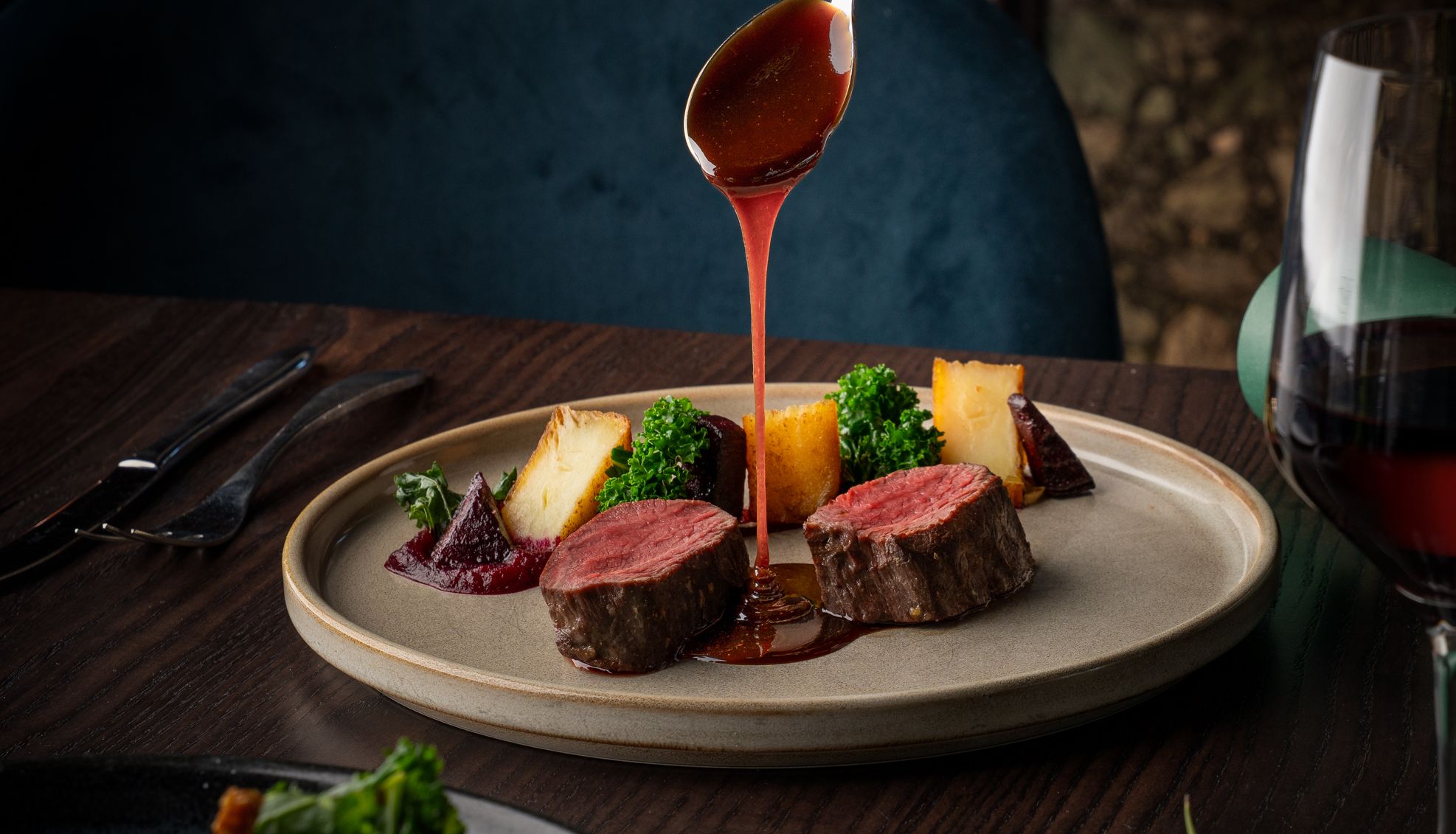 Hand pouring sauce over sliced beef tenderloin with vegetables and a glass of red wine on a dinner table.