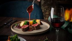 Hand pouring sauce over sliced beef tenderloin with vegetables and a glass of red wine on a dinner table.