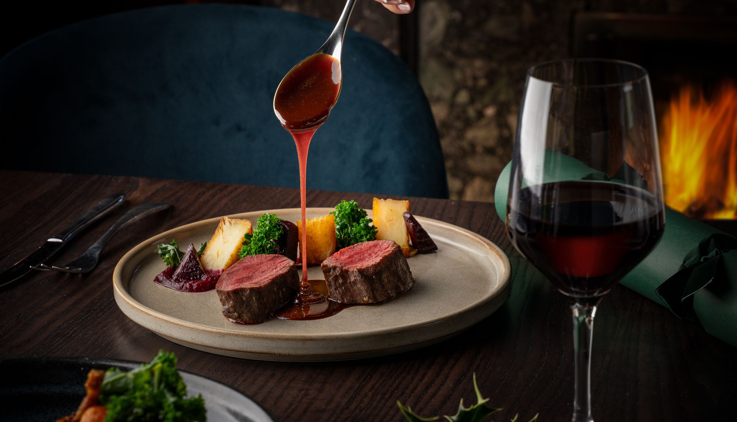 Hand pouring sauce over sliced beef tenderloin with vegetables and a glass of red wine on a dinner table.