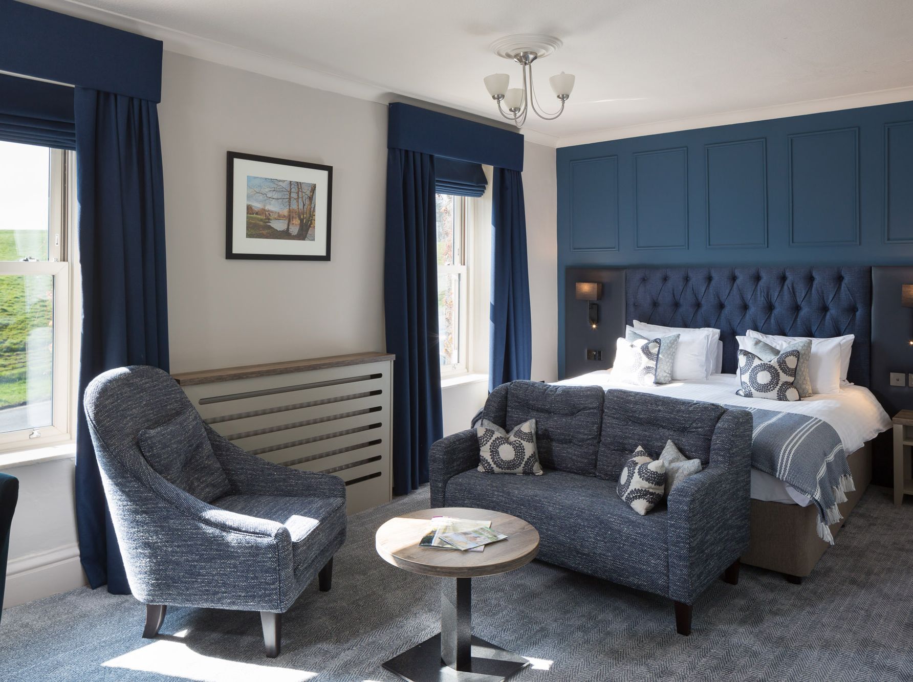Modern hotel room with blue and grey decor, featuring a large bed, armchair, sofa, and a small round table.